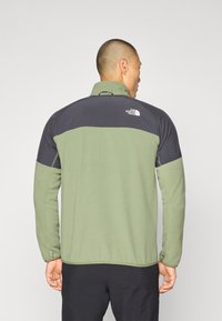 Zelena flis jakna s crnim gornjim dijelom. Ima visoki ovratnik, duge rukave i logo na leđima. Glatka tekstura, dizajnirana za outdoor nošenje.
