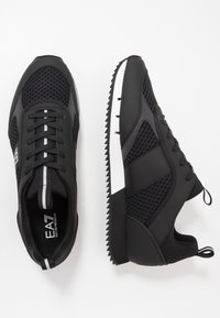 Zapatillas deportivas negras con paneles de malla, suelas de goma texturizadas y un acento blanco en el lateral. Cuentan con cordones y una pestaña de agarre; diseño elegante.