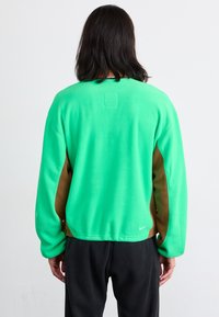 Zelena flis majica s smešnimi stranskimi akcenti, sproščen kroj, okrogli ovratnik in pravokotna obloga na hrbtu. Nike logo na obodu.