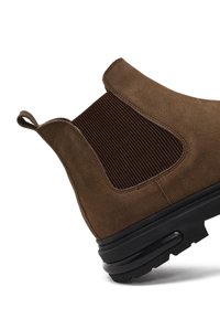 Stivale alla caviglia in suede marrone con pannello elastico a coste, linguetta e suola in gomma nera. Presenta una punta rotonda e una texture liscia.