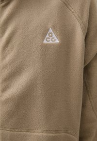 Fleece-Pullover in einem hellbraunen Farbton mit strukturiertem Oberflächen. Mit einem weißen gestickten Dreieck-Logo auf der Brust. Sichtbare Nahtdetails.