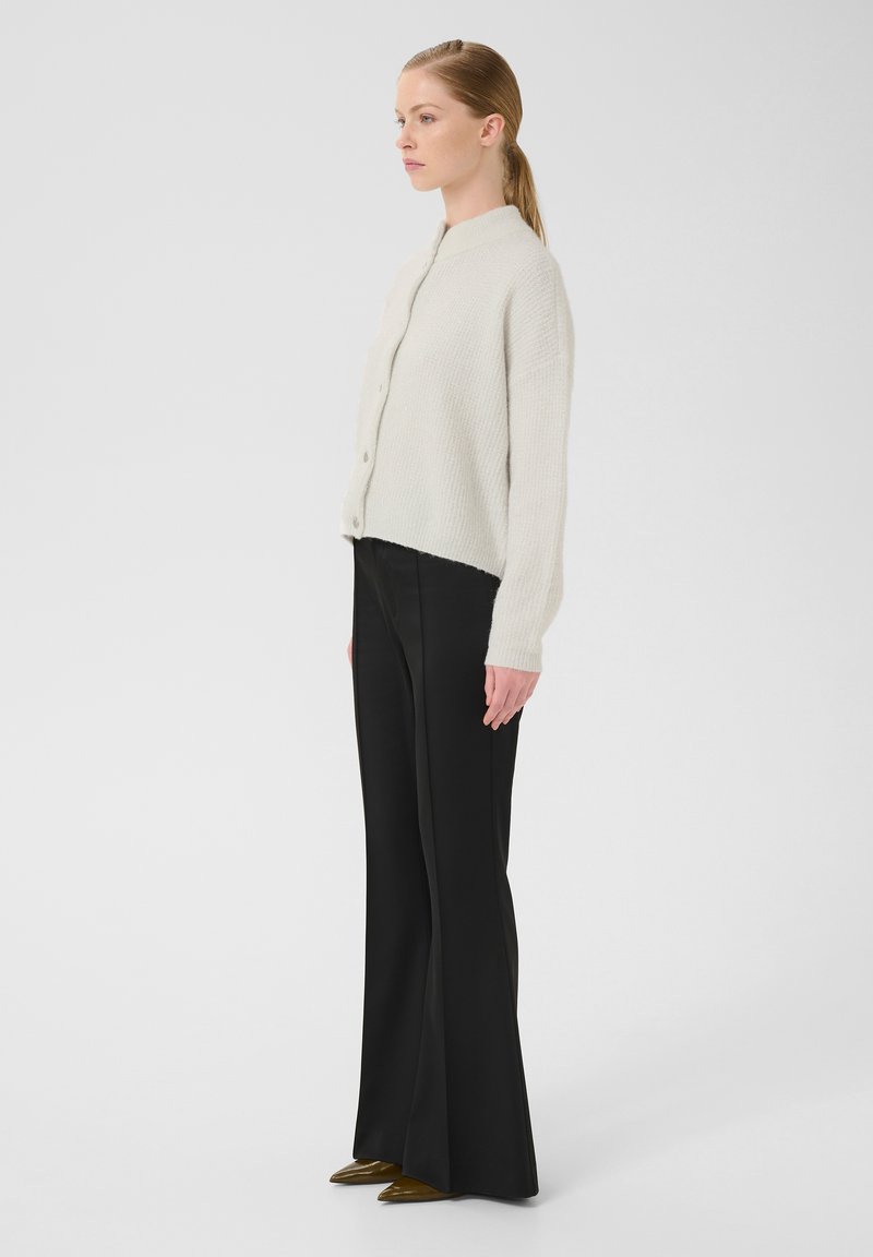 Cardigan surdimensionné en tricot crème avec un col haut, fermeture par boutons et texture côtelée, associé à un pantalon large noir et des chaussures vertes à bout pointu.