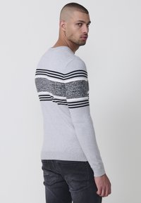 Suéter gris claro con un patrón texturizado de rayas horizontales en blanco y negro en el pecho y los brazos. Diseño ajustado con puños acanalados.