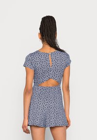 Macacão azul-marinho com padrão floral branco, mangas curtas, costas abertas com um recorte em forma de gota, e um corte relaxado com calções que terminam na metade da coxa.