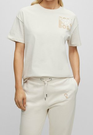 Woman wearing a cream-colored T-shirt with “BOSS” logo three times on chest and matching sweatpants with drawstring waist.