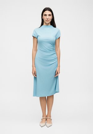 Femme debout, portant une robe midi bleu clair à manches courtes avec fronces latérales, associée à des chaussures blanches à bout pointu et bride, sur fond blanc.