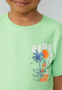 Lichtgroene katoenen T-shirt met een grafisch ontwerp van palmbomen, een zon en surfplanken. De tekst zegt "GROTE GOLF GROTE PLEZIER!" in felle kleuren.