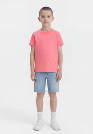 Junge steht, trägt ein korallenrotes kurzärmeliges Hemd, hellblaue Jeansshorts, weiße Socken und weiße Turnschuhe vor einem einfarbigen Hintergrund.