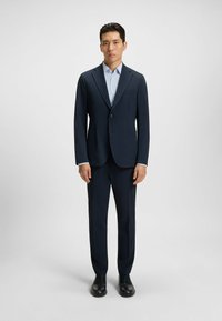 Marinblå kostym med kantskärningar, enknappsstängning och två framfickor. Ljusblå skjorta under. Svarta skor.