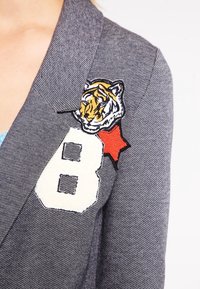 Šedý blazer s texturovanou tkaninou, zdobený vyšitou hlavou tygra a bílým číslem "8" s červenou hvězdou jako akcentem.