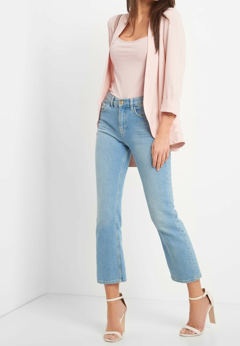 Femme portant un blazer rose clair, un haut rose pâle, un jean court bleu clair et des escarpins beige à bride à la cheville, debout devant un fond blanc.