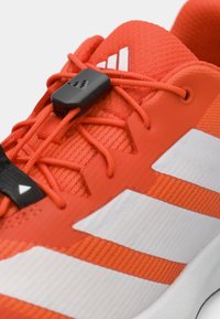 Κόκκινο αθλητικό παπούτσι με υφασμάτινη επάνω επιφάνεια, μαύρα κορδόνια, πλαστικό κλείδωμα κορδονιών και λευκές ρίγες στο πλάι. Λείο καουτσούκ πέλμα με πορτοκαλί λεπτομέρειες.