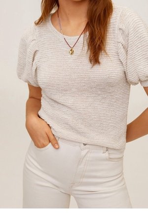 Kobieta ubrana w biały, teksturowany top z krótkimi, bufkami rękawami, białe spodnie z wysokim stanem oraz złoty wisiorek na czerwonym sznurku.