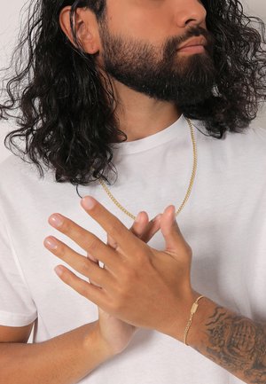 Goldene Kettenhalskette an einer Person, die ein weißes T-Shirt trägt. Die Hände sind gefaltet und zeigen ein passendes goldfarbenes Armband sowie ein Tattoo am Unterarm.