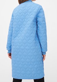 Ljusblå quiltad kappa med lång design, ribbade manschetter och ett diamantmönster över tyget, visad från baksidan.