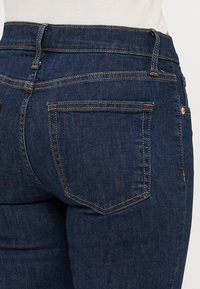 Jean en denim bleu foncé avec une taille haute, comportant deux poches arrière et des détails de surpiqûres contrastantes. Texture lisse et coupe ajustée.