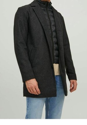 Homme portant un manteau en laine gris foncé superposé à une veste matelassée noire, un pull beige et un jean bleu clair sur un fond uni.