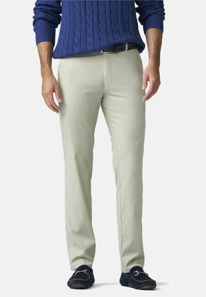 Homme portant un pantalon beige clair, une ceinture bleu marine, un pull en maille torsadée bleu marine et des mocassins bleu marine, debout devant un fond uni.