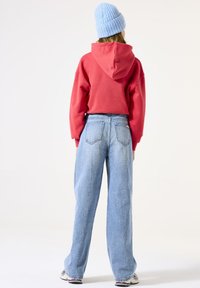 Persona vista desde atrás que lleva una sudadera roja con capucha, jeans de pierna ancha azul claro, zapatillas blancas y un gorro de punto azul claro sobre un fondo blanco.