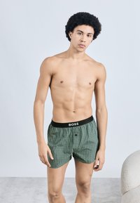 Zöld kockás boxeralsó fekete derékrésszel, amelyen a "BOSS" felirat látható. Puha anyag és kényelmes szabás. Modell világos háttér előtt áll.