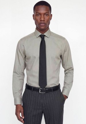 Homme portant une chemise rayée gris clair, une cravate noire, une ceinture noire et un pantalon à fines rayures gris foncé, debout sur fond blanc.