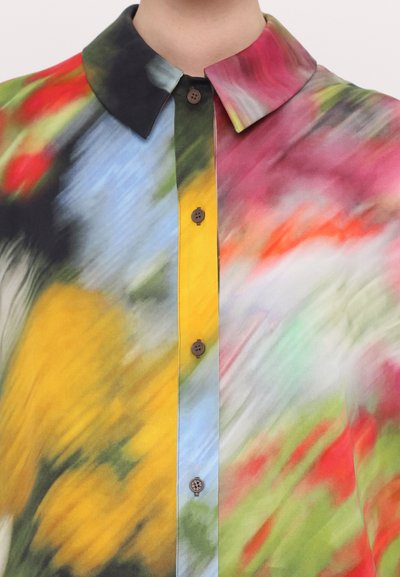 Blouse colorée à motifs abstraits avec un col, présentant des motifs tourbillonnants en rouge, jaune, bleu et vert, fermée par des boutons marron.