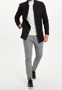 Zwarte, met kraag, buitenjas met knopenfront. Witte gebreide trui daaronder. Grijze op maat gemaakte broek met zijnaad. Zwarte sneakers maken de look af.