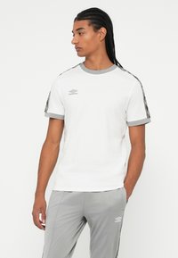 Mann med flettet hår iført hvite og grå Umbro sportsklær, stående med den ene hånden i lommen, vendt mot venstre, mot en enkel hvit bakgrunn.