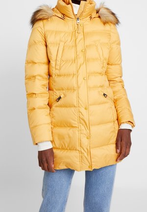 Manteau d’hiver matelassé jaune avec capuche bordée de fourrure, fermeture éclair frontale et boutons-pression, porté sur un jean bleu clair et des manches de pull blanches.