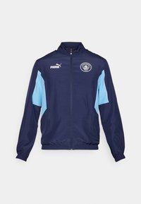 Temno modra športna jakna z svetlo modrimi detajli, poln zadrgo, s stoječim ovratnikom, elastičnimi manšetami ter logotipom Manchester City na prsih.