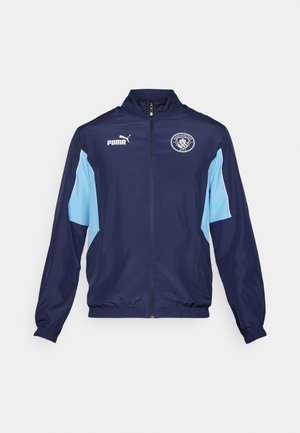 Marineblauwe sportjas met lichtblauwe accenten, volledige rits, staande kraag, elastische manchetten en met het Manchester City-logo op de borst.