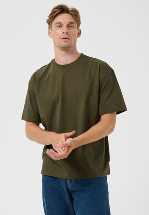 Joven con camiseta verde oliva y vaqueros azules de pie frente a un fondo blanco liso.