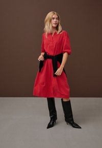 Femeie în picioare, purtând o rochie midi roșie cu mâneci bufante, centură neagră legată în jurul taliei și cizme negre înalte, cu vârf ascuțit, pe un podea gri.