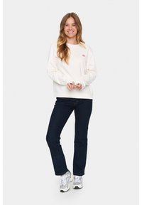 Felpa bianca con scollo rotondo, caratterizzata da un piccolo logo rosa, abbinata a jeans dritti blu scuro e sneakers grigie.