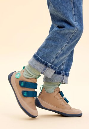 Child wearing tan leather sneakers with blue straps and green socks, standing on tiptoes with rolled-up blue jeans against a neutral background.