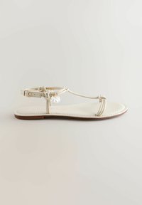 Lapos bézs szandál vékony pántokkal, bokapántos csattal és kis fehér díszítőmedálokkal az oldalpánton.