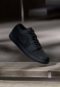 Μαύρα αθλητικά παπούτσια Nike με άνω μέρος από λείο δέρμα και σουέτ, σκούρες κορδόνες, τρυπητό μπροστινό μέρος και λεπτομέρεια με κόκκινη ραφή. Σχεδίαση επίπεδης σόλας.