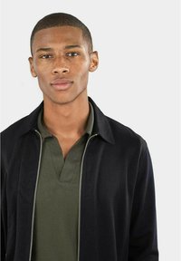 Schwarze zipfelbare Jacke mit Kragen über einem dunkelgrünen Poloshirt. Der Stoff wirkt glatt mit einem figurnahen Design. Schlichter, moderner Stil.