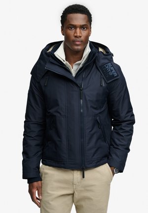 Mężczyzna nosi granatową bluzę z kapturem na zamek błyskawiczny z logo "Superdry" na ramieniu, na białej koszuli i beżowych spodniach.