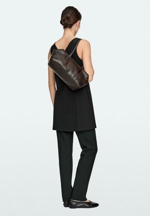 Femme aux cheveux attachés en chignon, portant une tunique noire sans manches, un pantalon noir, des ballerines noires, portant un sac à main en cuir marron sur l'épaule, vue de dos.