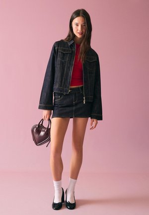 Jonge vrouw met een donkeren spijkerjack en rok, rood shirt, witte sokken, zwarte hakken, die een handtas in de vorm van een hart vasthoudt tegen een roze achtergrond.