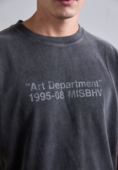 Nahaufnahme einer Person, die ein verwaschenes schwarzes T-Shirt mit weißem Text "Art Department 1995-08 MISBHV" auf der Brust trägt.