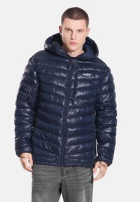 Granatowa pikowana kurtka z błyszczącym wykończeniem, zapinana na zamek błyskawiczny z przodu, z kapturem. Posiada pikowaną fakturę i małe logo na piersi.