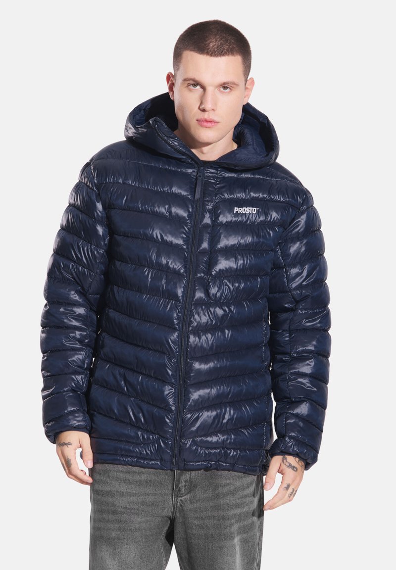 Granatowa pikowana kurtka z błyszczącym wykończeniem, zapinana na zamek błyskawiczny z przodu, z kapturem. Posiada pikowaną fakturę i małe logo na piersi.