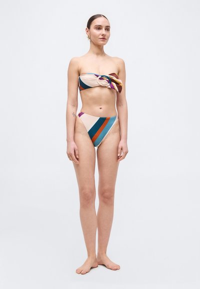 Vrouw staat blootsvoets en draagt een strapless, meerkleurige gestreepte bikini met broekje met hoge taille, tegen een eenvoudige lichte achtergrond.
