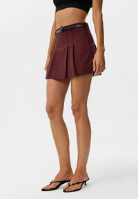 Bordeauxrode geplooide rok met een aangesloten taille en zwarte riem, met een gladde stofstructuur en een uitlopende zoom. Draagbaar met zwarte teenslippers.