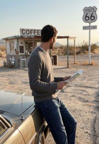 Ein Mann in Pullover und Jeans lehnt an einem Auto, hält eine Karte in der Nähe eines Kaffee-Standes und eines Route 66-Schildes in einer Wüstenlandschaft.