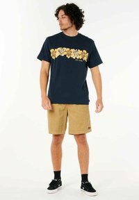 Navyblaues T-Shirt mit gelbem Blumenmuster auf der Brust, kombiniert mit beigen Cordshorts und schwarzen Turnschuhen mit weißen Sohlen.