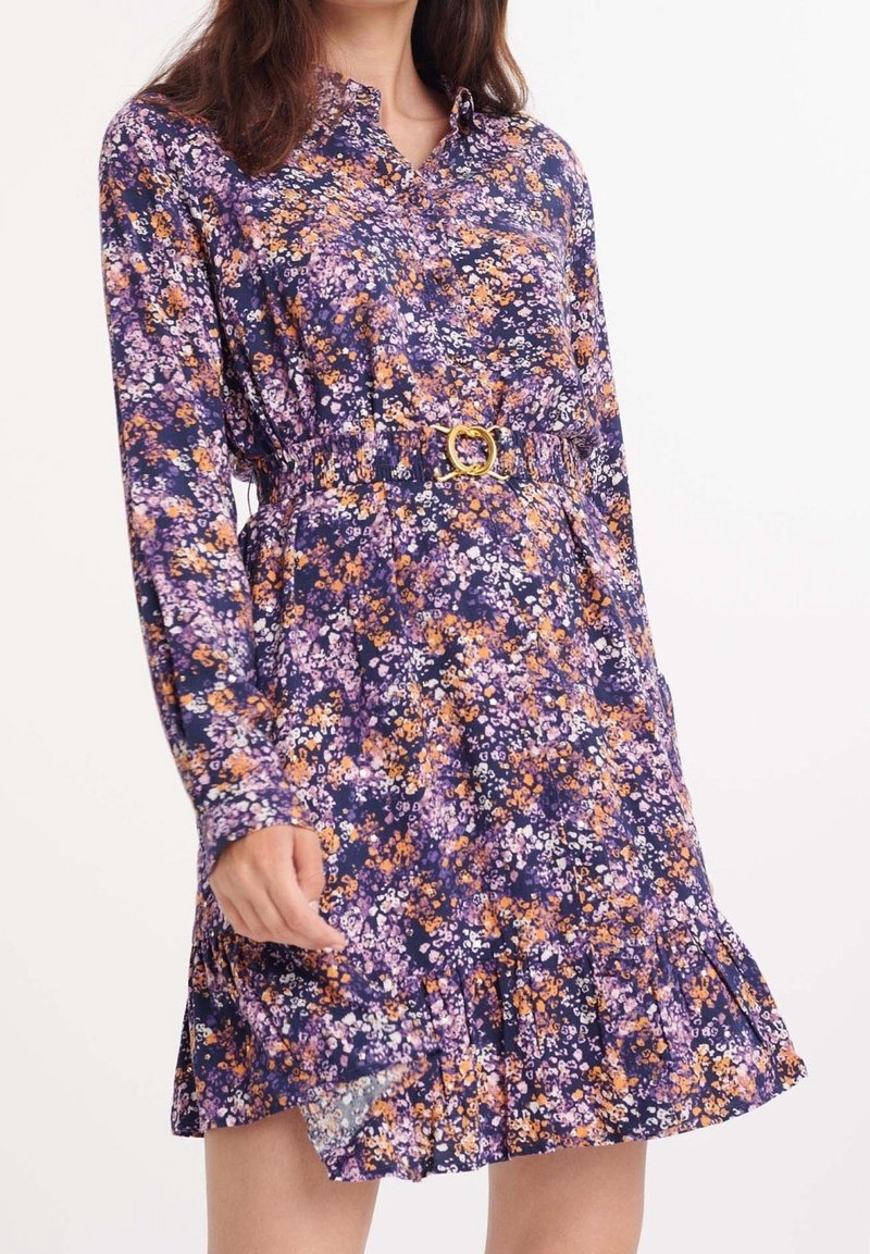 Abito floreale di colore viola scuro con fiori arancioni e bianchi, maniche lunghe, colletto, vita affusolata con una fibbia dorata e orlo arricciato.