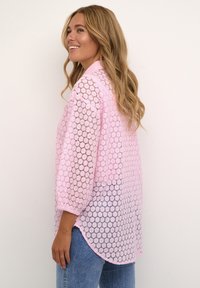 Light pink lace blouse with a round pattern, three-quarter sleeves, and a curved hem, layered over a casual top.
